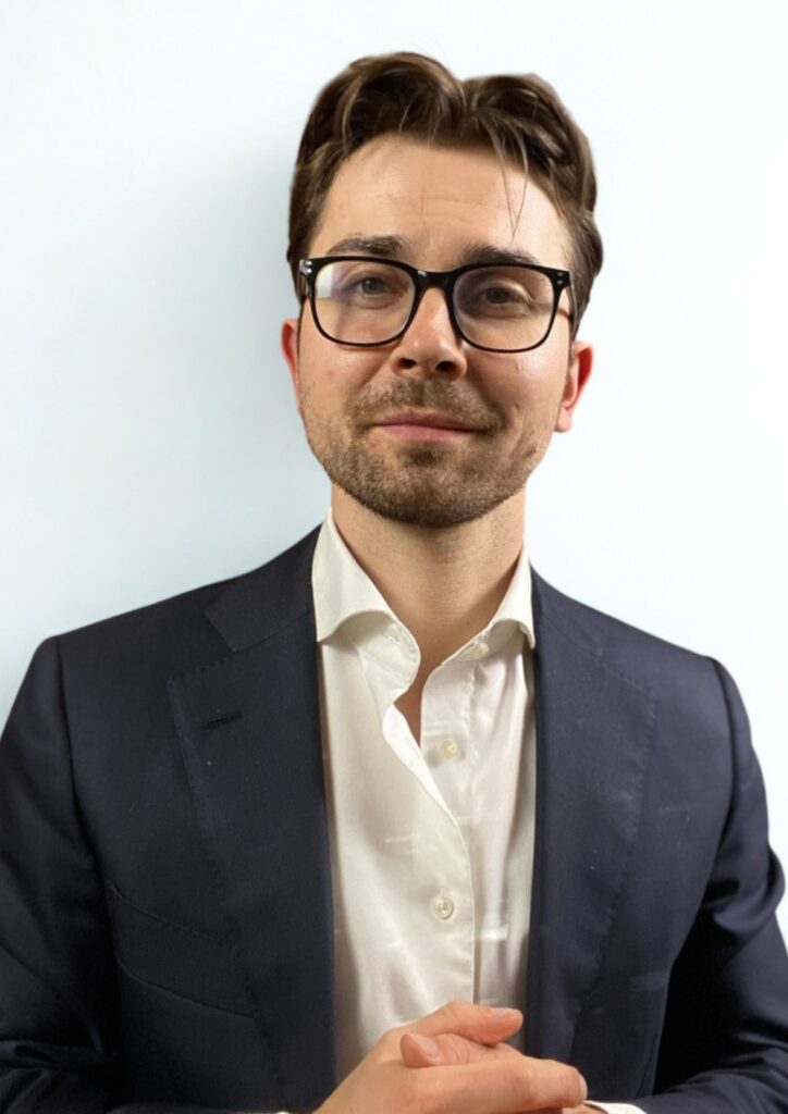 Simon Schreiner Portrait, trägt ein weißes Hemd und ein dunkel blaues sakko. Er ist 26 Jahre alt, mittel lange haare, eckige brille, selbst bewusste haltung mit leichten lächeln